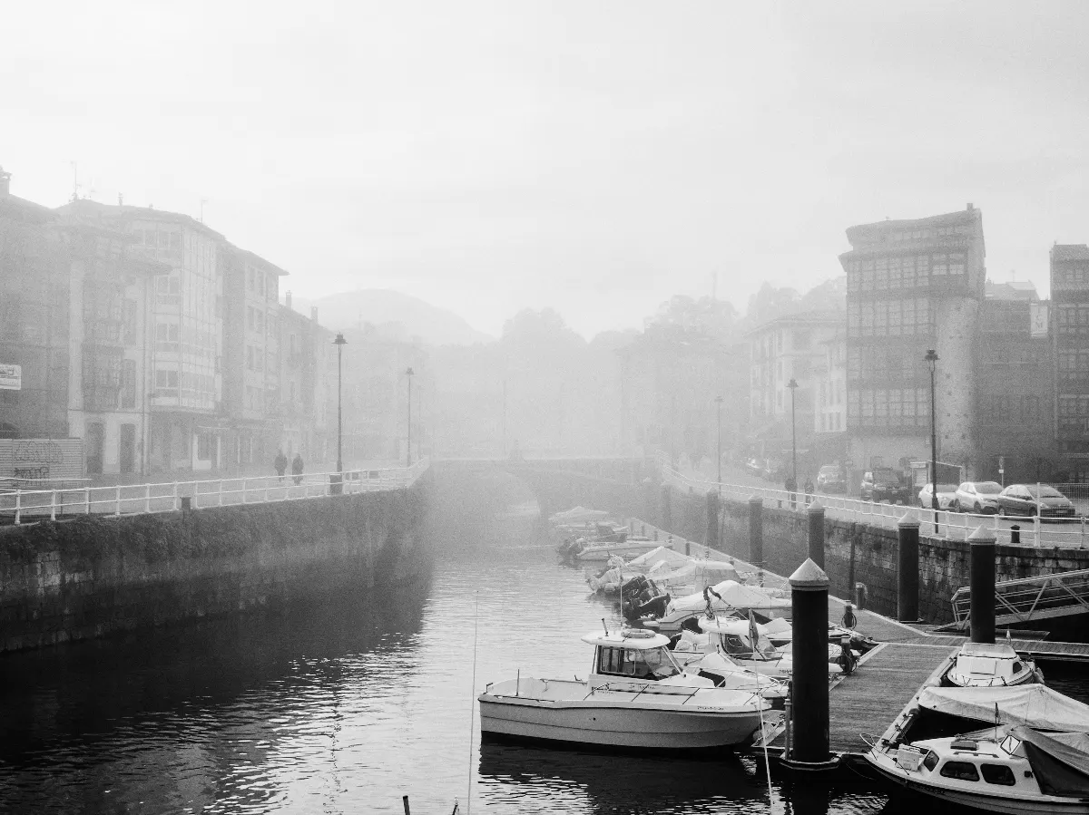 Foggy riverside scene with small boats docked along a pier, historic buildings lining both sides, and a bridge in the background, all enveloped in mist.